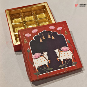 A red and gold boxed food product shown open to reveal gold tray compartments inside, with the lid leaning in front. The lid features a decorative illustration of two white cows with pink lotus flowers under a black arched panel, on a light gray background.
