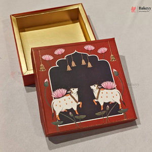 A square decorative red and gold gift box shown open with its lid placed in front. The lid features a dark central arch with two white spotted cows, pink lotus flowers, hanging bells, and green leaves, set against a light gray background.