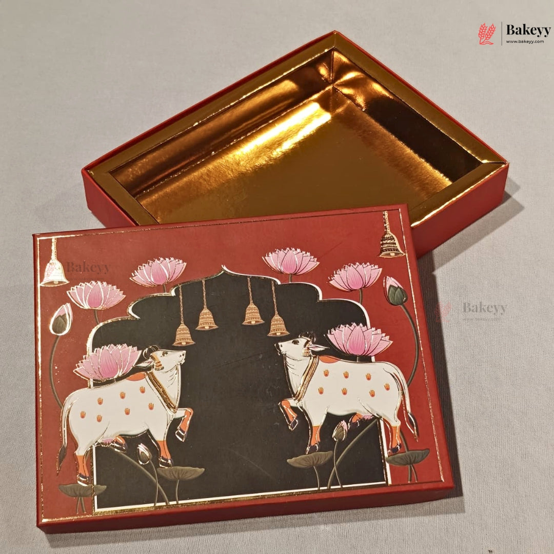 A decorative rectangular gift box for sweets shown open, with a red exterior and a shiny gold interior. The lid features a black central panel with two white cows decorated with pink lotus flowers, gold bells, and small green leaves, all on a red border.