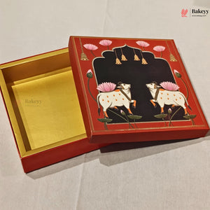 An open decorative red and gold bakery box with a painted lid showing two white spotted cows under pink lotus flowers on a black background, photographed on a light fabric surface.