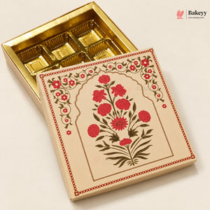 An open gold-colored rectangular box with six compartments, shown next to its illustrated cream lid featuring red flowers and green leaves, on a light beige background. A Bakeyy logo is visible in the top right corner.