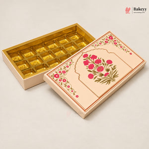 An open beige gift box with gold foil compartments, shown next to its decorated lid featuring pink floral patterns and green leaves on a light beige background.