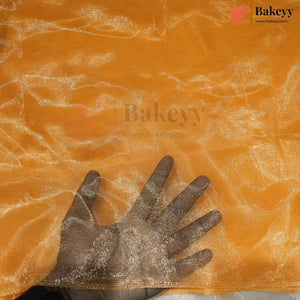 A close-up of a sheer, shimmering orange mesh fabric with a hand placed underneath to demonstrate its transparency and texture. The Bakeyy brand logo is visible in the top right corner.