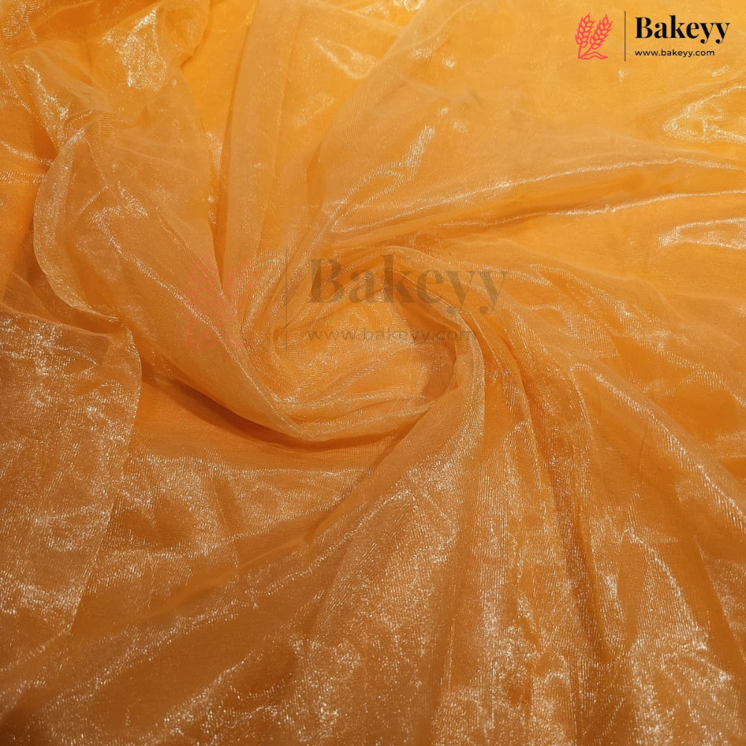 A close-up of sheer, shimmering orange tulle or organza fabric, swirled and draped to show its texture and reflective quality. The Bakeyy brand logo is visible in the top right corner.