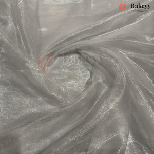 Shimmering silver organza fabric with a metallic reflective finish, draped in a swirl pattern. A Bakeyy brand logo is visible in the top right corner.
