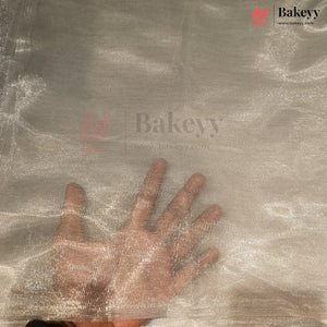 A hand is placed underneath a sheer, shimmering metallic fabric to show its texture and transparency. The fabric has a silver-gray sheen and is branded with Bakeyy watermarks.