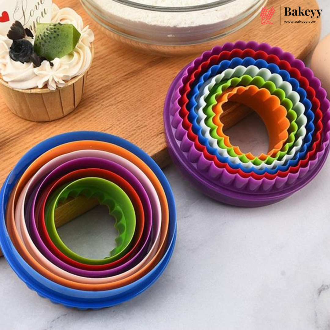 A set of colorful round cookie cutters in nested rings, shown on a light marble surface with a cupcake and a bowl of flour in the background. The cutters are shown in two stacks: one on blue, orange, and purple, and another on purple, red, green, blue, orange, and white.
