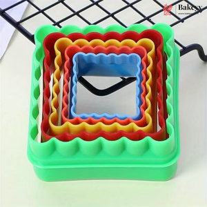 A set of nested square cookie cutters in green, red, yellow, and blue, arranged on a light surface with a black wire rack in the background. A BakeCy brand logo is visible in the top right corner.