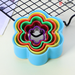 A set of nested flower-shaped cookie cutters in rainbow colors, including blue, red, green, yellow, and purple, with a metal scoring blade visible in the center. The cutters are arranged on a light surface with a black wire cooling rack in the background.