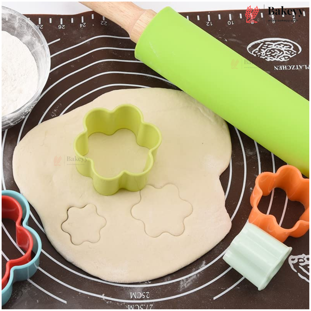 A baking scene with a light green flower-shaped cookie cutter placed on rolled-out dough, alongside an orange heart-shaped cutter, a turquoise heart-shaped cutter, a bright green rolling pin, and a brown silicone baking mat with measurement markings.