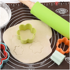 A baking scene with a light green flower-shaped cookie cutter placed on rolled-out dough, alongside an orange heart-shaped cutter, a turquoise heart-shaped cutter, a bright green rolling pin, and a brown silicone baking mat with measurement markings.