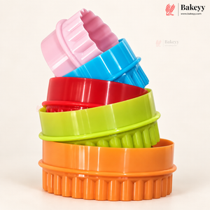 A stack of five colorful round baking pans with fluted edges in pink, blue, red, green, and orange, photographed on a clean white background. The Bakeyy logo is visible in the top right corner.