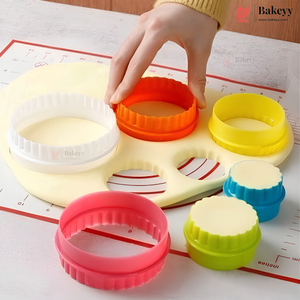 A set of colorful round cookie cutters and stamp molds arranged on a white surface with a baking mat. A hand is pressing down an orange circular cutter, which is part of a larger pale yellow silicone tray.