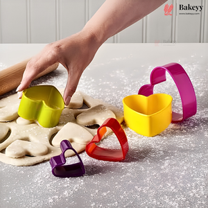 A set of colorful heart-shaped cookie cutters in yellow, red, purple, and magenta, shown on a floured baking surface with dough and a hand reaching in. A rolling pin is also visible, and the Bakeyy brand logo appears in the top right corner.