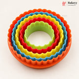 A set of five nested round scalloped cookie cutters in rainbow colors: orange, yellow, red, blue, and green, arranged on a clean white background. The Bakeyy brand logo is visible in the top right corner.
