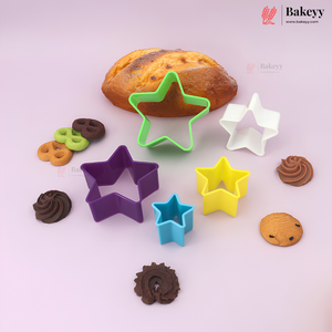 Five star-shaped cookie cutters in purple, green, white, yellow, and blue, displayed with various baked cookies and a loaf of bread on a light pink background. The Bakeyy brand logo is visible in the top right corner.