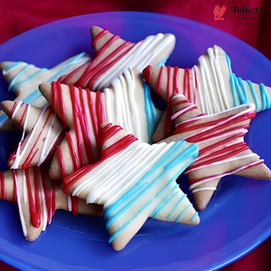 Star-shaped sugar cookies decorated with red, white, and blue drizzle, arranged on a blue plate. A Bakey logo watermark is visible in the top right corner.