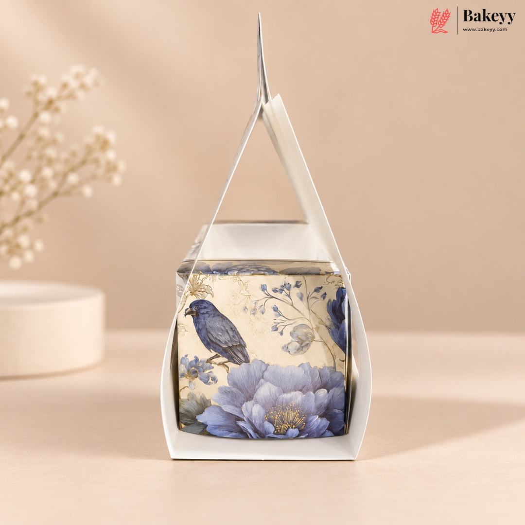 A square white gift box with a clear window and triangular handle, decorated with a vintage-style illustration of a blue bird perched on large lavender-blue flowers against a cream background. The box is photographed on a warm beige surface with soft floral props blurred in the background.