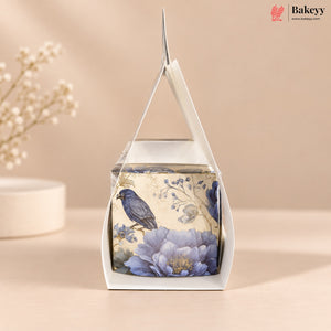 A square white gift box with a clear window and triangular handle, decorated with a vintage-style illustration of a blue bird perched on large lavender-blue flowers against a cream background. The box is photographed on a warm beige surface with soft floral props blurred in the background.