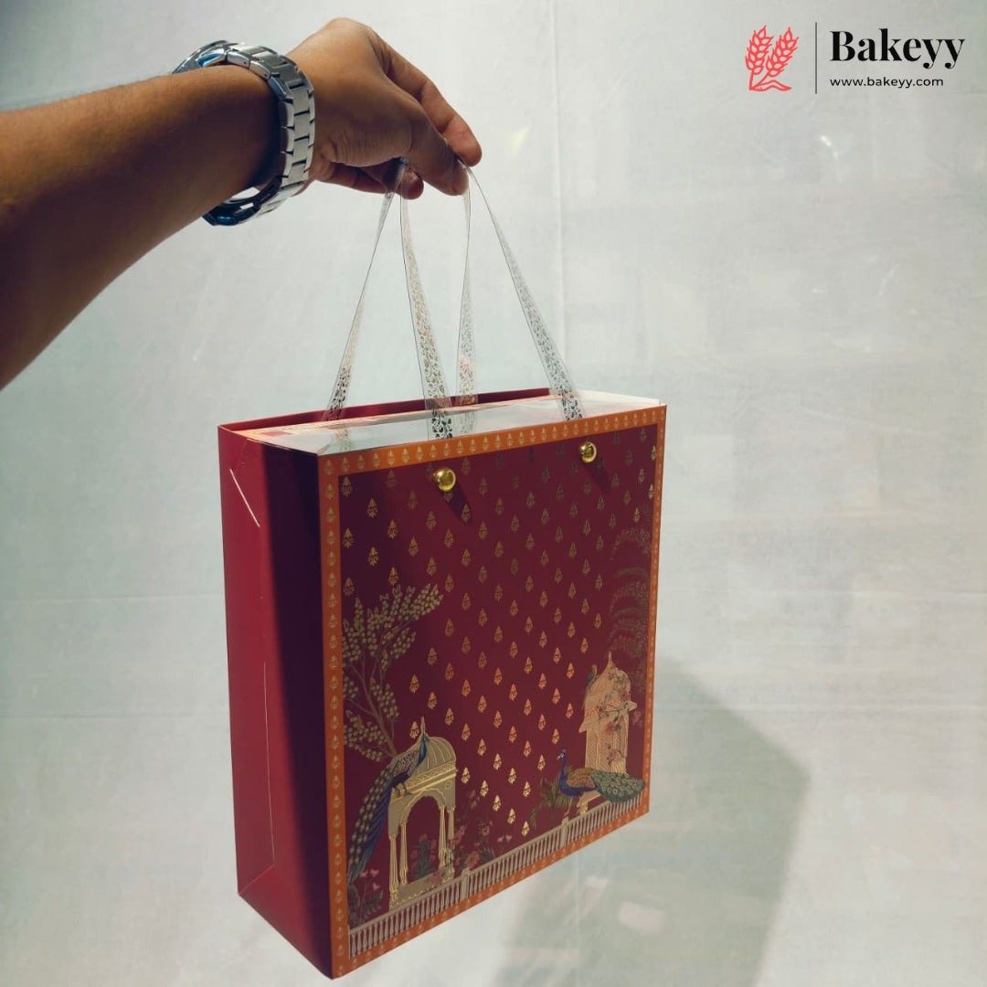 A hand holds a red gift bag with gold decorative patterns and white handles against a light gray background. The Bakeyy logo is visible in the top right corner.