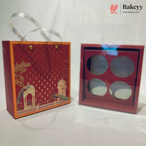 A red gift box set with a decorative printed paper bag featuring peacock and architectural motifs, alongside a matching square box with four circular cutouts and a clear lid, shown on a white fabric background. A Bakeyy logo watermark is visible in the top right.