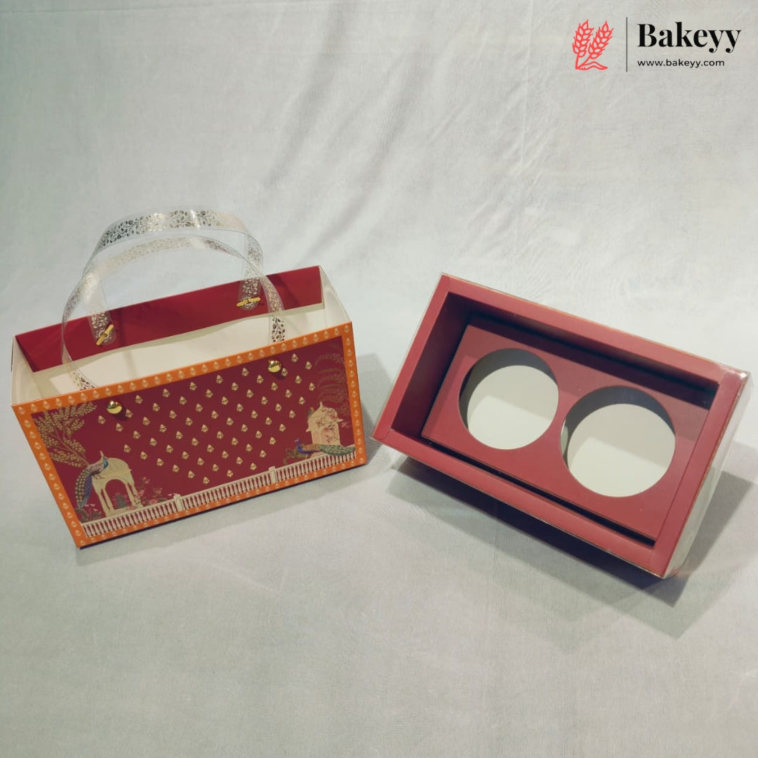 A red and orange bakery gift box set with a decorative printed exterior and a matching interior tray featuring two circular cutouts, photographed on a light gray fabric backdrop. A Bakeyy logo watermark is visible in the top right corner.