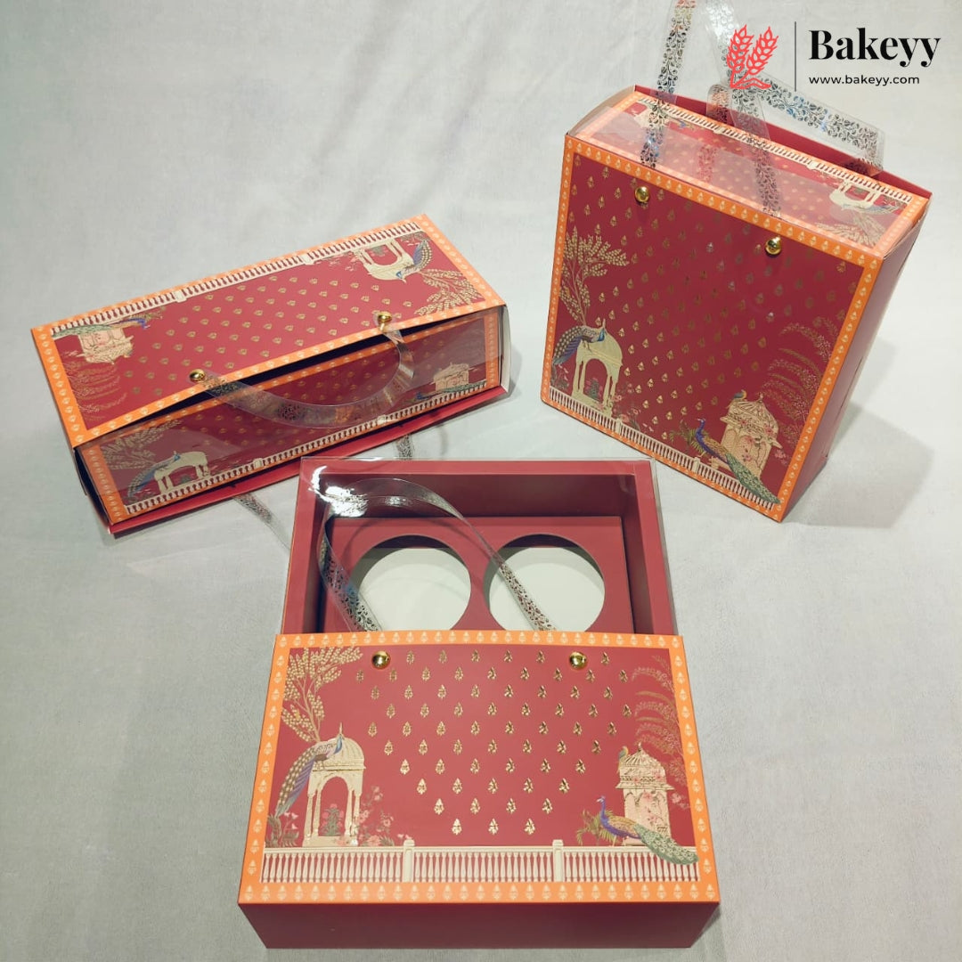 Three red and orange decorative gift boxes with illustrated architectural designs and a clear plastic top, shown on a light gray fabric background. One box is open, revealing two round white inserts, and the Bakeyy logo appears in the top right corner.