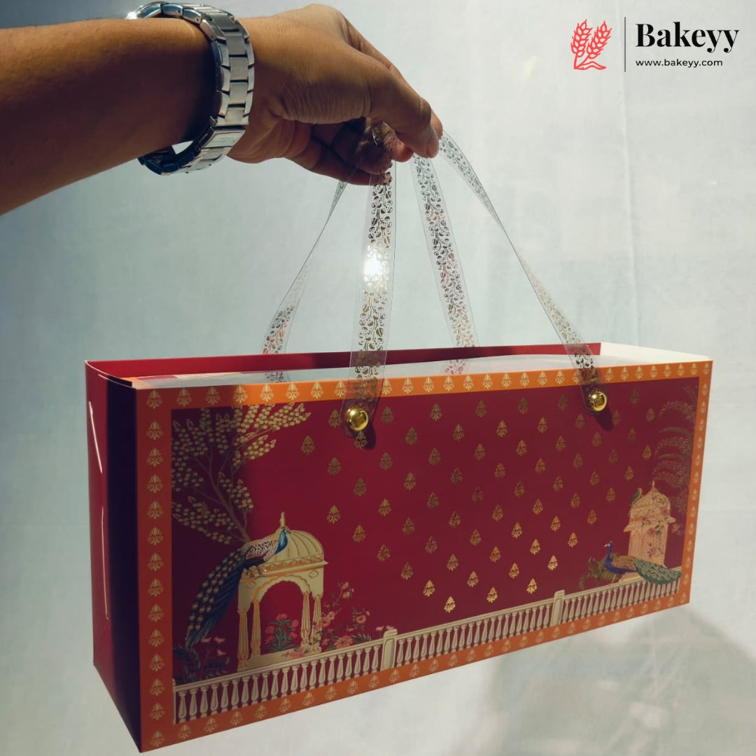 A hand holds a rectangular red gift bag with gold decorative patterns, illustrated peacocks, and clear patterned handles. The Bakeyy logo is visible in the top right corner.
