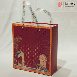 A decorative red bakery gift box with gold foil patterns, peacock illustrations, and clear plastic handles, photographed on a white fabric background. The Bakeyy logo is visible in the top right corner.