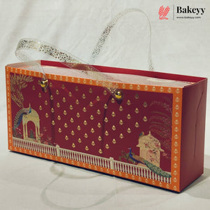 A rectangular red bakery gift box with a transparent handle, decorated with gold motifs, peacock illustrations, and a white border. The Bakeyy logo is visible in the top right corner.