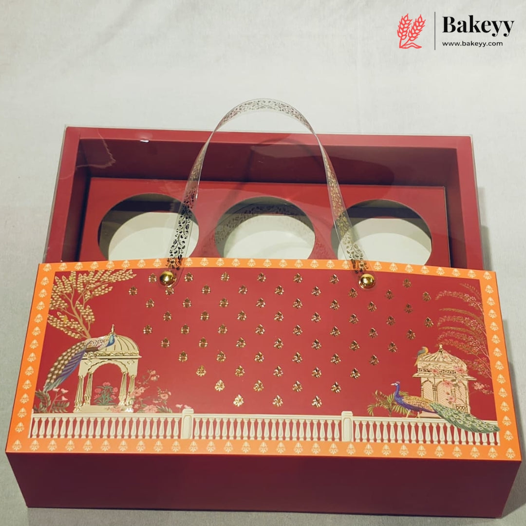 A red gift box with a hinged lid, featuring a decorative front panel with gold foil motifs, peacocks, and a white railing. Inside the open box are three round white plates with clear plastic handles.