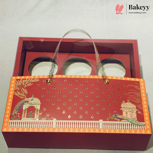 A red gift box with a hinged lid, featuring a decorative front panel with gold foil motifs, peacocks, and a white railing. Inside the open box are three round white plates with clear plastic handles.