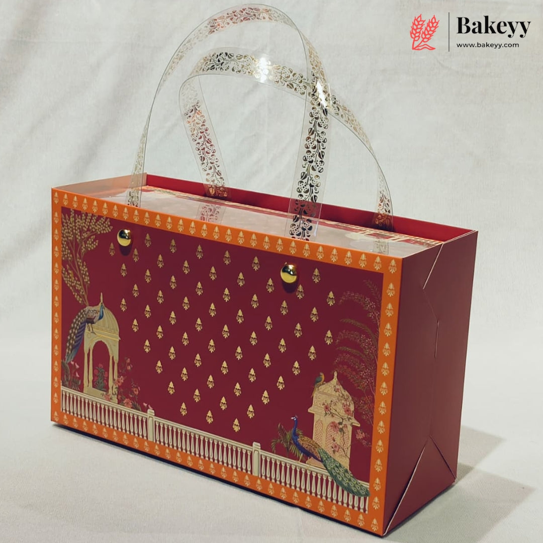 A decorative rectangular bakery gift box with clear plastic handles, featuring a deep red front panel with gold motifs, an orange border, and peacock illustrations, photographed on a light neutral background.