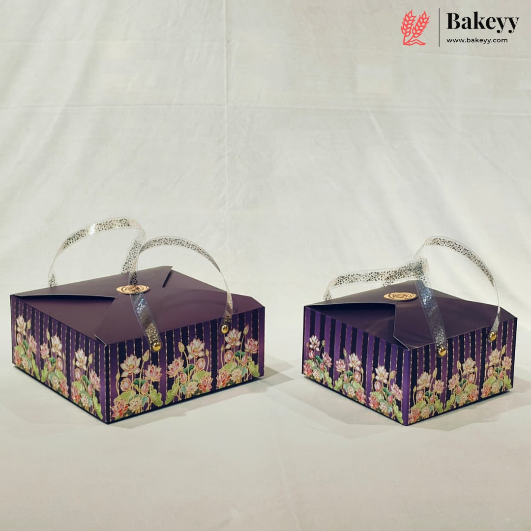 Two rectangular bakery gift boxes with purple floral patterns and silver handles, displayed on a white fabric backdrop. The Bakeyy logo is visible in the top right corner.