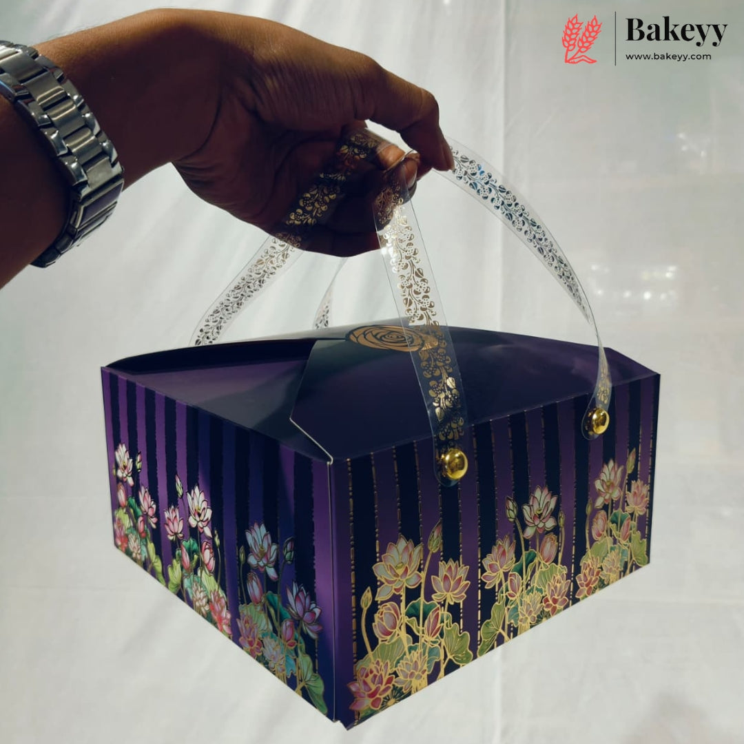 A hand holds a square purple bakery box with floral lotus designs and clear plastic handles. The box features vertical purple stripes and a gold seal on top.
