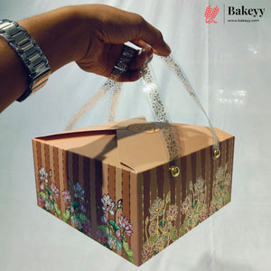 A rectangular brown bakery gift box with floral printed designs on the sides, clear plastic handles, and a hand holding it. The Bakeyy logo is visible in the top right corner.