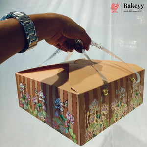 A hand wearing a silver watch holds a decorative cardboard bakery box with floral illustrations and gold-tone hardware against a white backdrop. The Bakeyy logo appears in the top right corner.