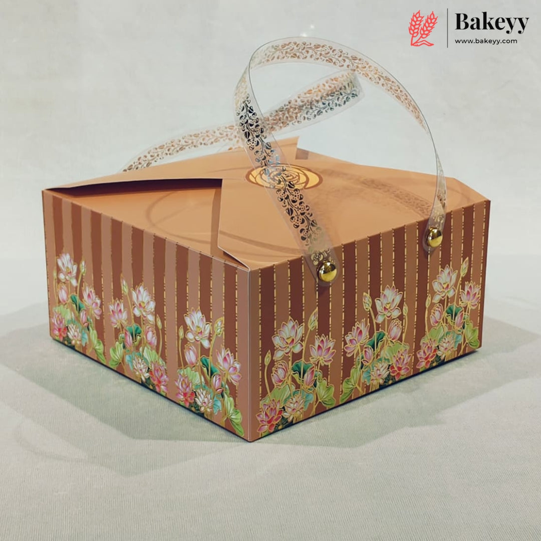 A square bakery gift box with a brown and gold striped pattern, decorated with pink lotus flowers along the bottom. The box features a clear plastic handle with a black lace pattern and small gold bells at the ends, set against a neutral light gray background.