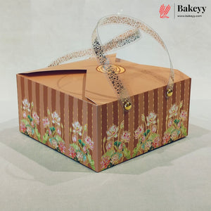 A square bakery gift box with a brown and gold striped pattern, decorated with pink lotus flowers along the bottom. The box features a clear plastic handle with a black lace pattern and small gold bells at the ends, set against a neutral light gray background.