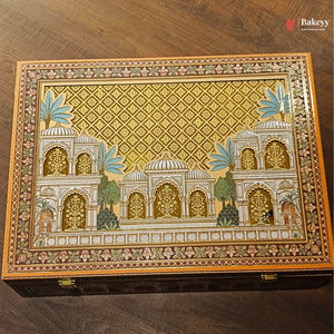 An ornate rectangular decorative box with a hand-painted Indian palace motif on the lid, featuring intricate gold, beige, and blue details, placed on a dark wooden surface.