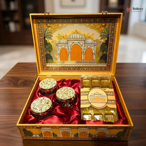 An ornate open gift box containing three small jars and a wrapped box of chocolates, set on a wooden table. The box has red satin lining and an illustrated lid showing a domed architectural scene.