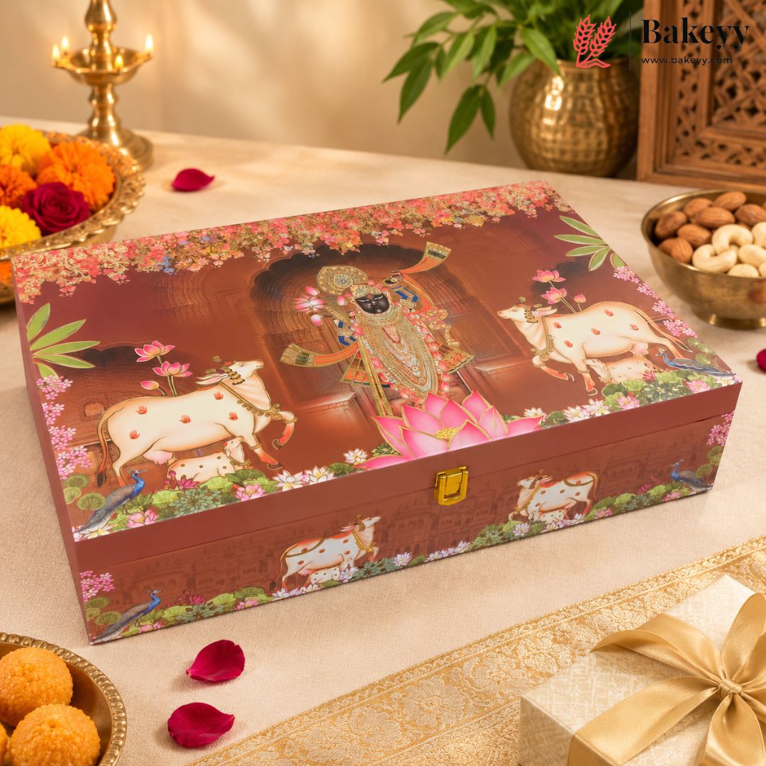 A decorative rectangular gift box with a reddish-brown background and colorful religious artwork featuring a seated figure, flowers, peacocks, and white cows. The box is styled on a beige tablecloth with candles, flowers, nuts, and a wrapped gift nearby.