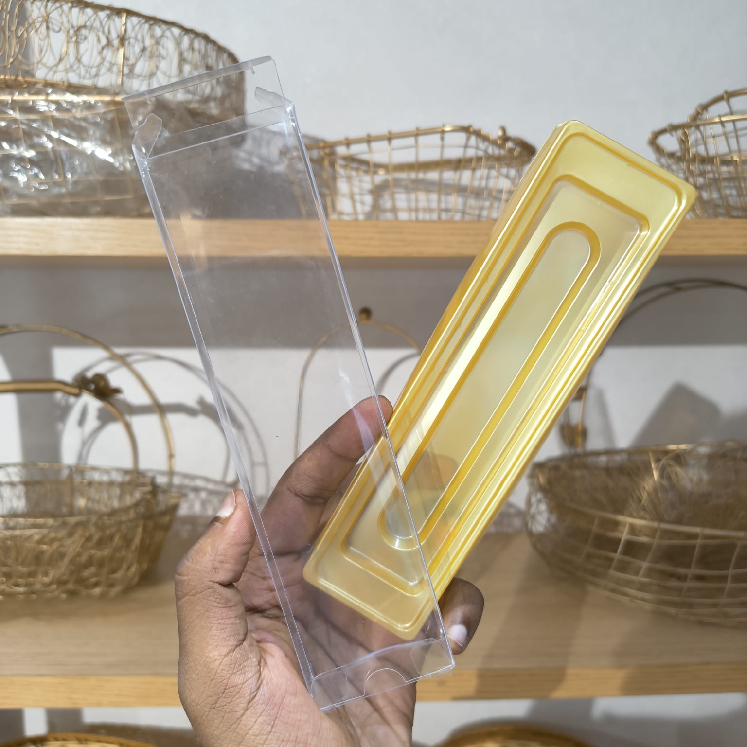 A hand holding a clear plastic rectangular box next to a translucent yellow plastic insert tray, with wire baskets on wooden shelves in the background.