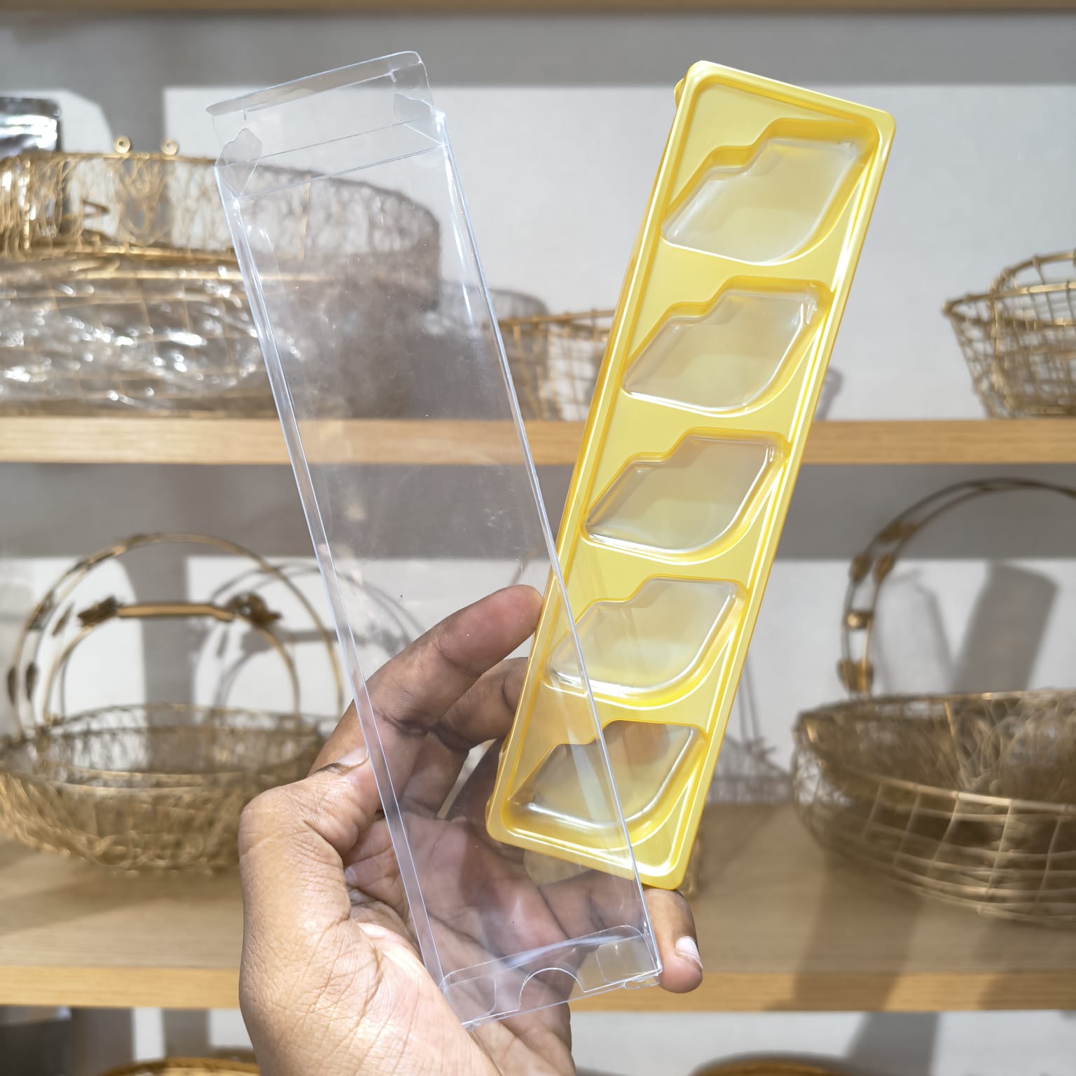 A hand holds a clear plastic rectangular box with four recessed compartments next to a yellow plastic tray with matching compartment shapes, against a blurred background of shelves with decorative baskets.