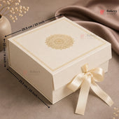 An off-white square bakery gift box with a gold mandala design on the lid, a cream satin ribbon bow, and black dimension arrows showing measurements of 25.5 cm by 10 cm by 25.