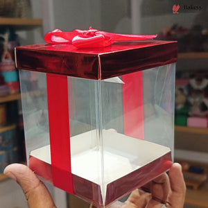 Clear gift box with red ribbon held by a hand, blurred indoor background