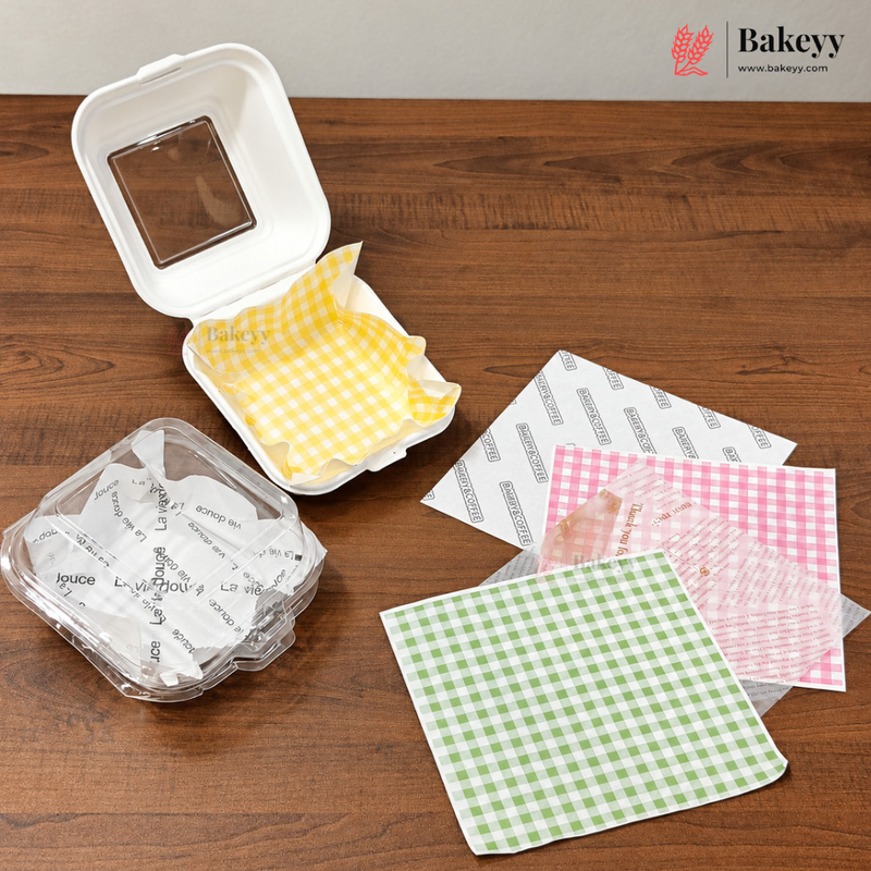 Bakeyy bakery packaging set on a wooden table, including a white styrofoam container with a clear window, a clear plastic container with printed liners, and several gingham paper liners in yellow, pink, and green.
