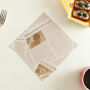 A square paper napkin printed with a vintage newspaper design featuring sepia text and images, styled on a white tabletop with a fork on the left and a slice of toast with blueberries in a yellow checkered dish on the top right.
