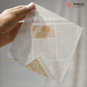 A hand holds a square piece of bakery gift wrap printed with elegant serif text reading 'Thank you for the lovely tea party' and a small illustration of a teacup and saucer. The wrap has a warm beige background with brown text and a decorative corner panel.