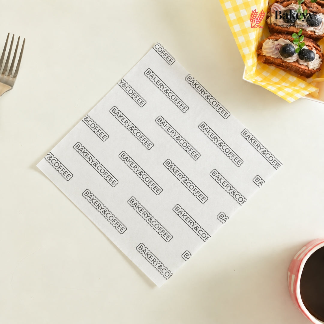 White paper napkins printed with the brand name 'Bakery & Coffee' repeated in black rectangular outlines, arranged on a light beige tabletop with a fork, blueberry tarts, and a cup of coffee nearby.